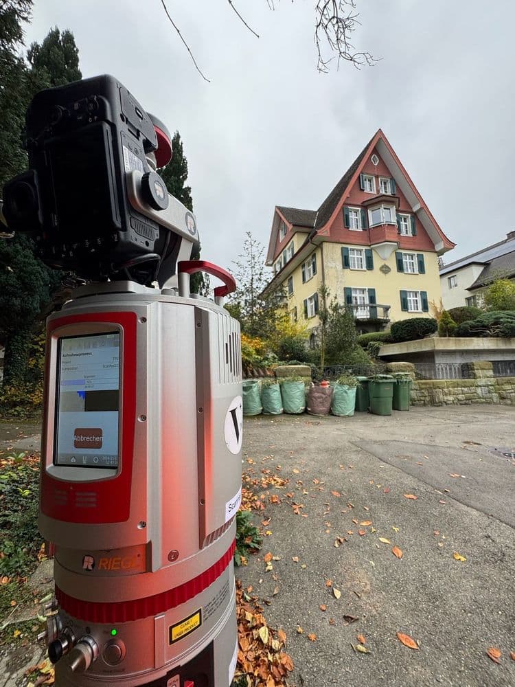 Ein Laserscanner steht auf einer Einfahrt vor einem Haus mit rotem Giebel und Herbstlaub. Im Hintergrund sind Mülltonnen und herbstliche Bäume zu sehen.