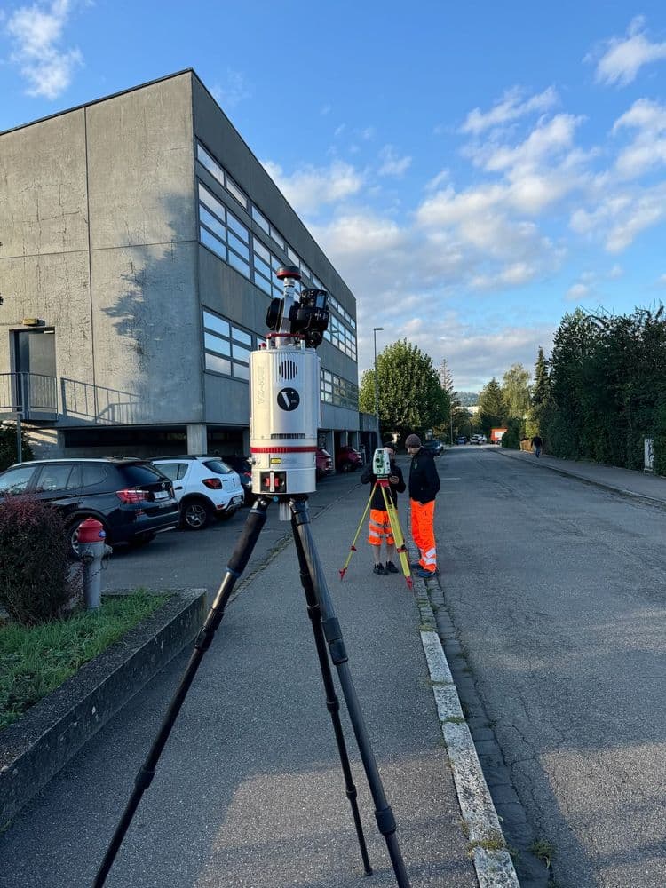Kameraausrüstung auf hohem Stativ wird von einem Techniker in Warnkleidung neben einem Gebäude bedient.