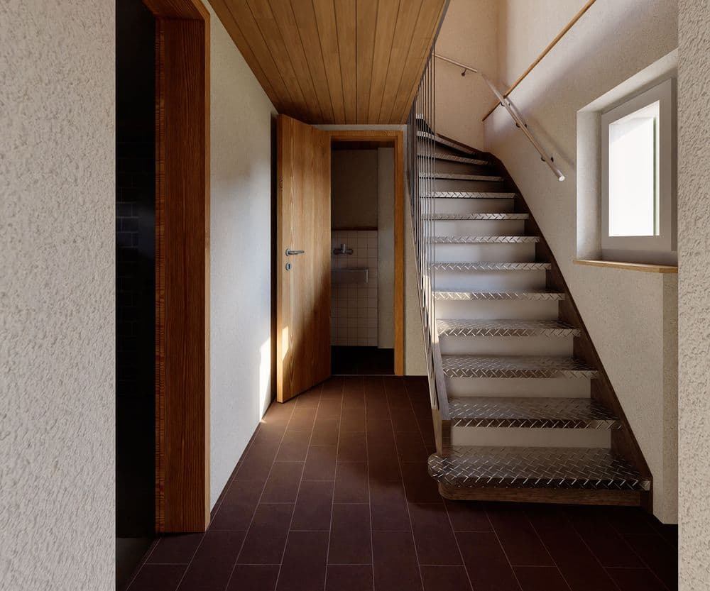 Innenansicht eines Treppenhauses mit Metalltreppe, weiß verputzten Wänden, Holzdecke, Fensterlicht und geöffneter Holztür zu einem kleinen Raum; brauner Fliesenboden und Geländer.