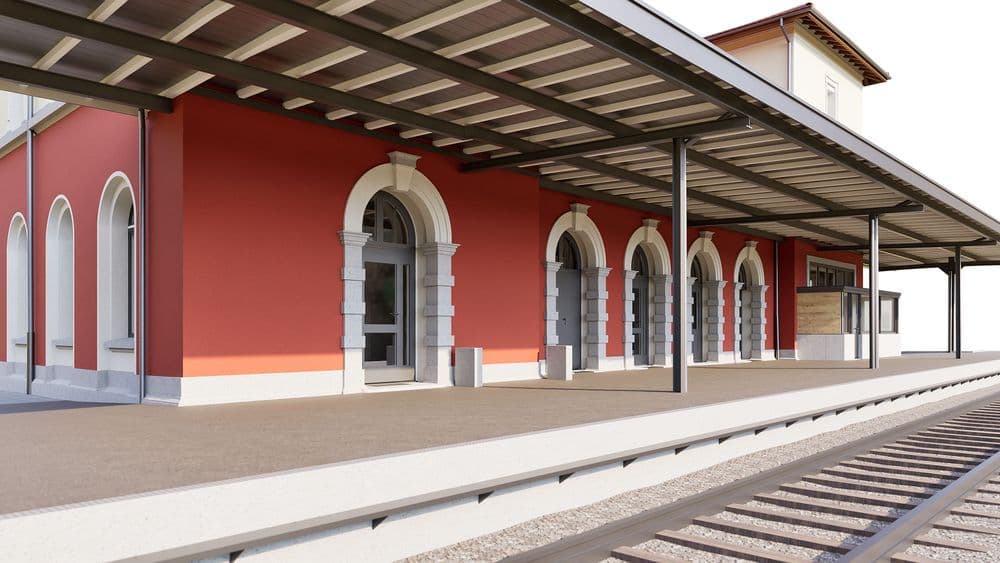 Bahnsteig eines historischen Bahnhofs mit roter Fassade, Rundbogenfenstern und überdachtem Perron; im Vordergrund Gleise, klare Tagesansicht.
