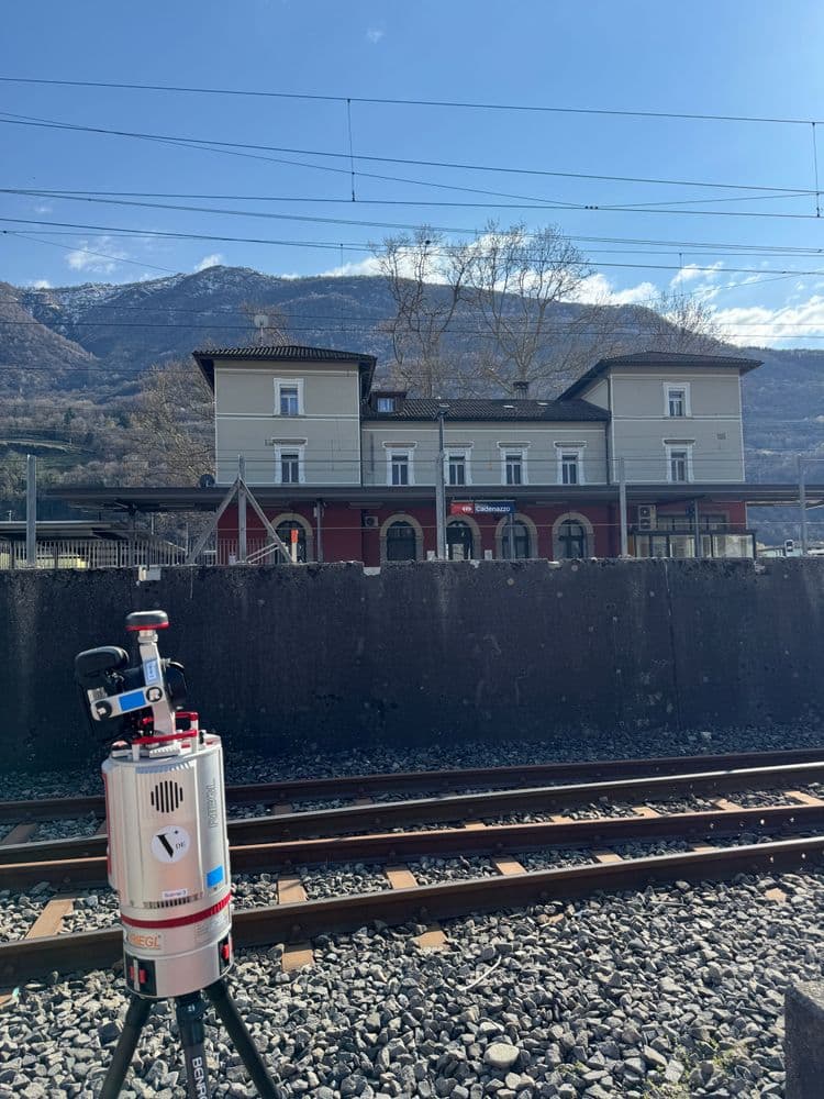 Kleiner Vermessungsroboter auf Stativ neben Bahngleisen, dahinter Bahnhof mit Oberleitungen und Alpenpanorama unter blauem Himmel.