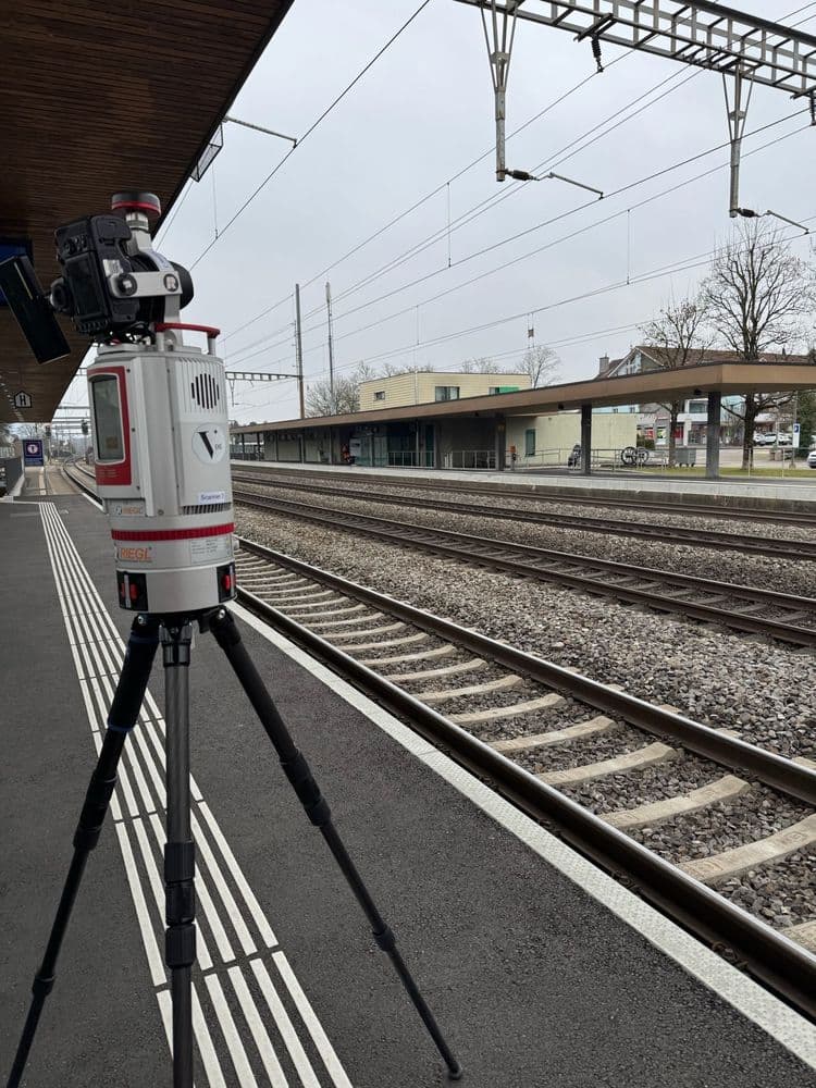 Ein großes Vermessungsgerät steht auf einem Stativ am Bahnsteig eines Bahnhofs, neben Bahngleisen. Der Himmel ist bewölkt und die Umgebung wirkt ruhig.