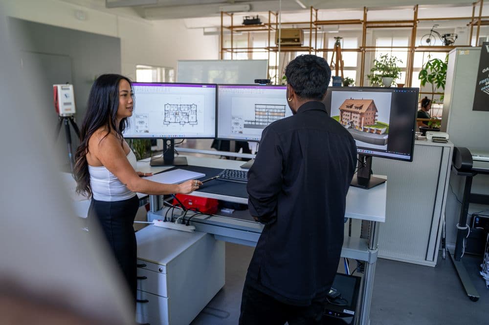 Büro mit zwei Personen, die an Schreibtischen mit mehreren Monitoren CAD-Software nutzen.