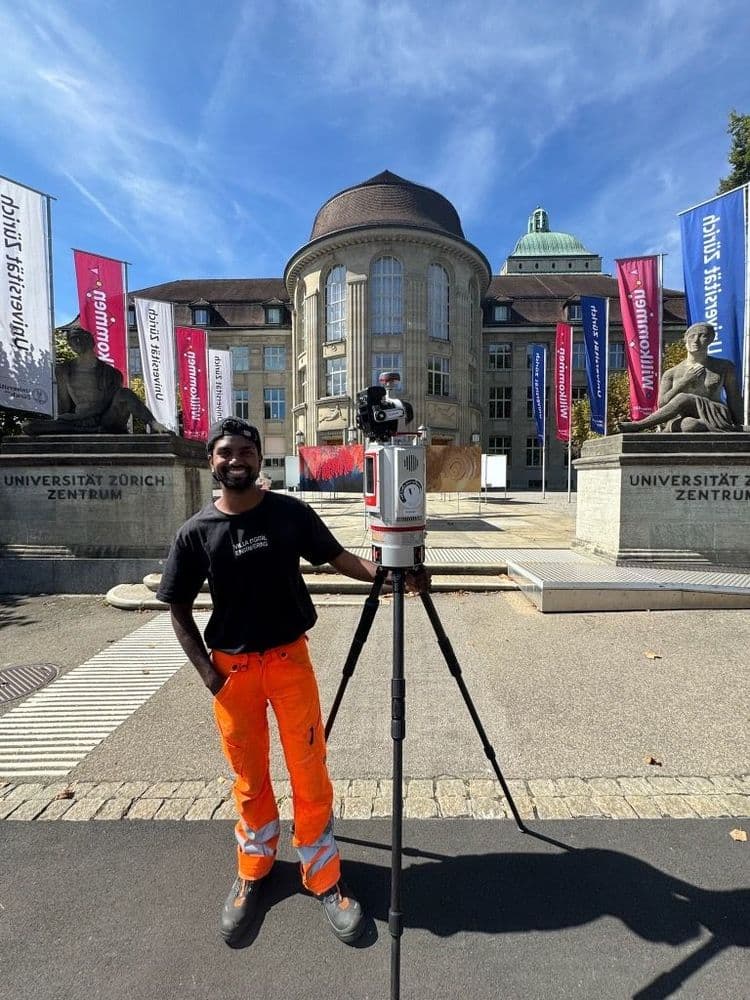 Vermessungsingenieur mit Stativ und 3D-Laserscanner vor der Universität Zürich Zentrum, blauem Himmel und Fahnen; Gebäude und Löwenskulpturen im Hintergrund.