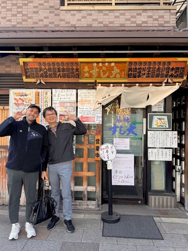 Zwei Personen stehen vor einem japanischen Restaurant. Das Gebäude hat ein auffälliges, mit Schriftzeichen versehenes Banner und mehrere Aushänge an der Wand.