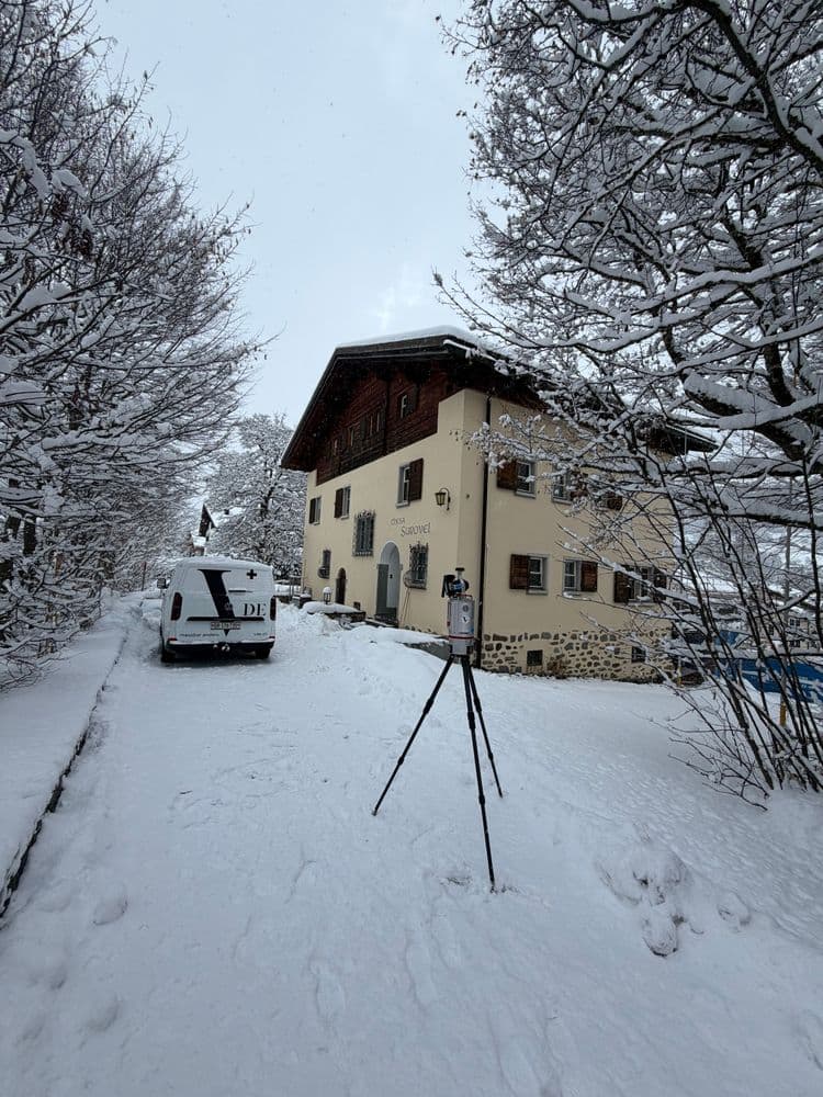 Verschneites Landhaus in Winterlandschaft mit schneebedeckten Bäumen, Einfahrt und geparktem Lieferwagen; vor dem Gebäude steht ein Stativ im Schnee.