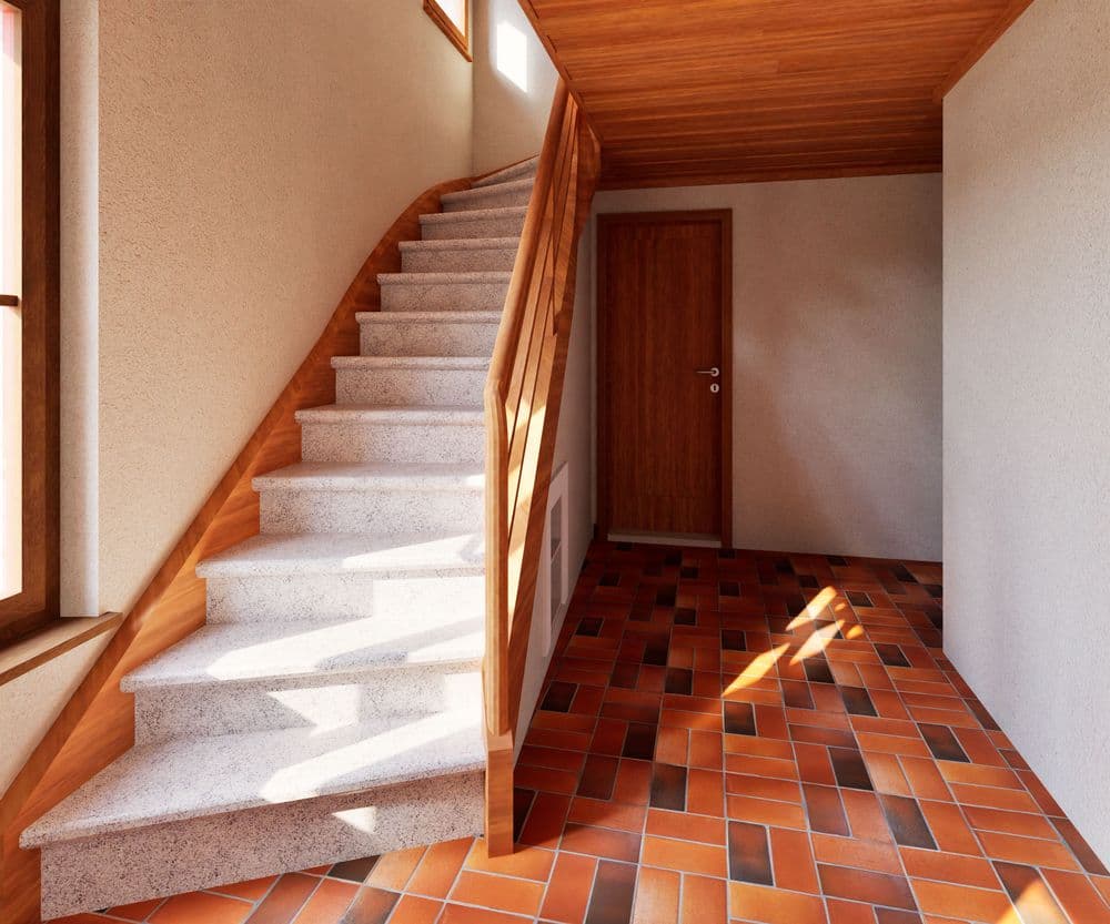 Heller Hausflur mit Treppe aus Stein, Holzgeländer und Holzdecke, Terrakottafliesenboden, Eingangstür und Fenster mit Sonnenlicht.