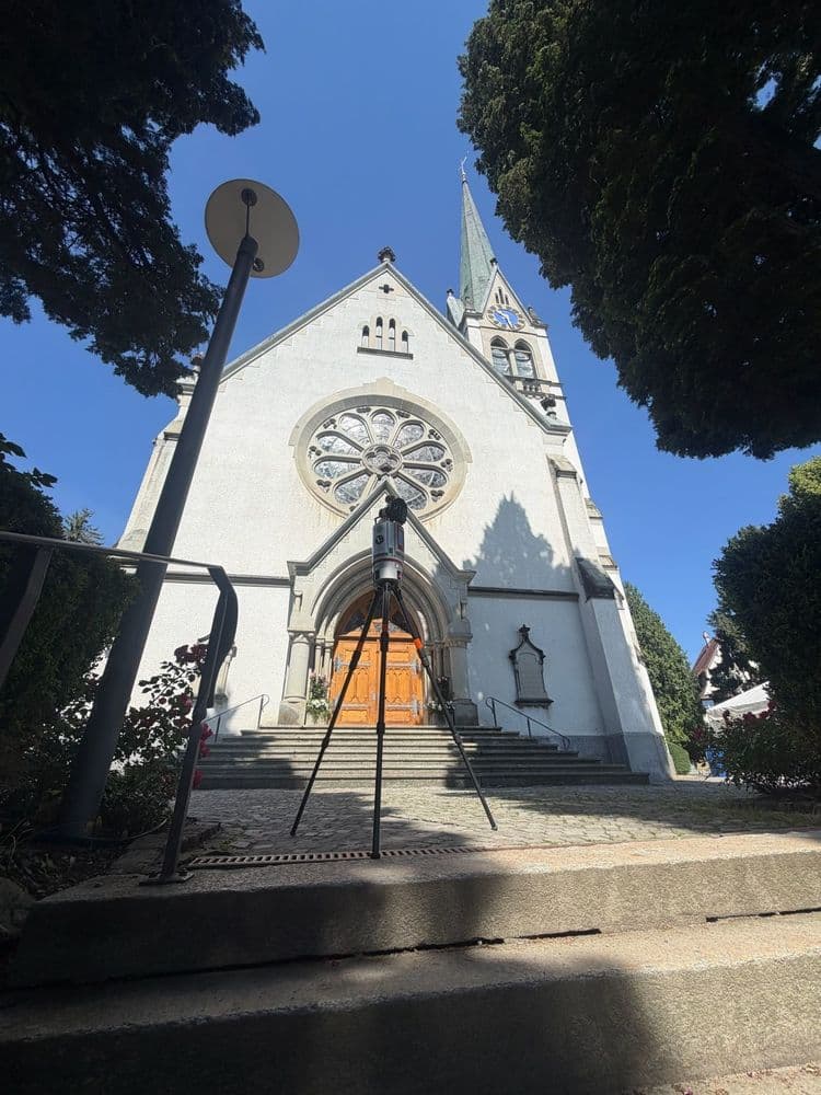 Frontansicht einer weißen Kirche mit Spitzenturm und Rosettenfenster, Treppe zum Holztor, aufgenommen aus Froschperspektive zwischen Bäumen bei blauem Himmel.