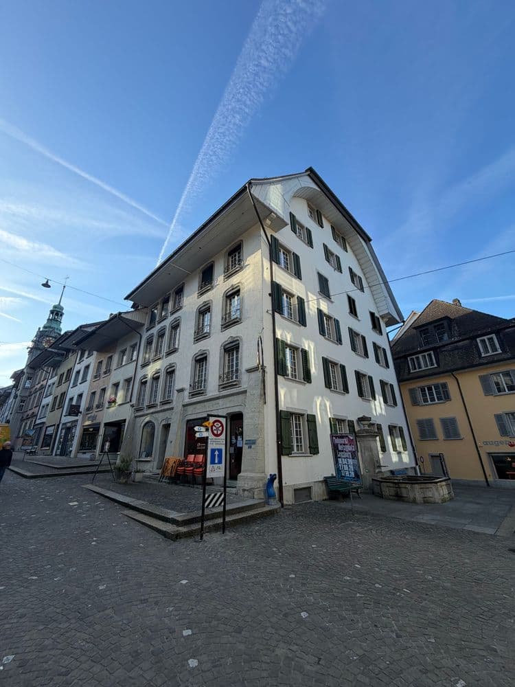 Historisches Altstadtgebäude an gepflastertem Platz mit Straßenschildern und blauem Himmel; perspektivische Weitwinkelaufnahme mit umliegenden Häusern und Kirchturm im Hintergrund.