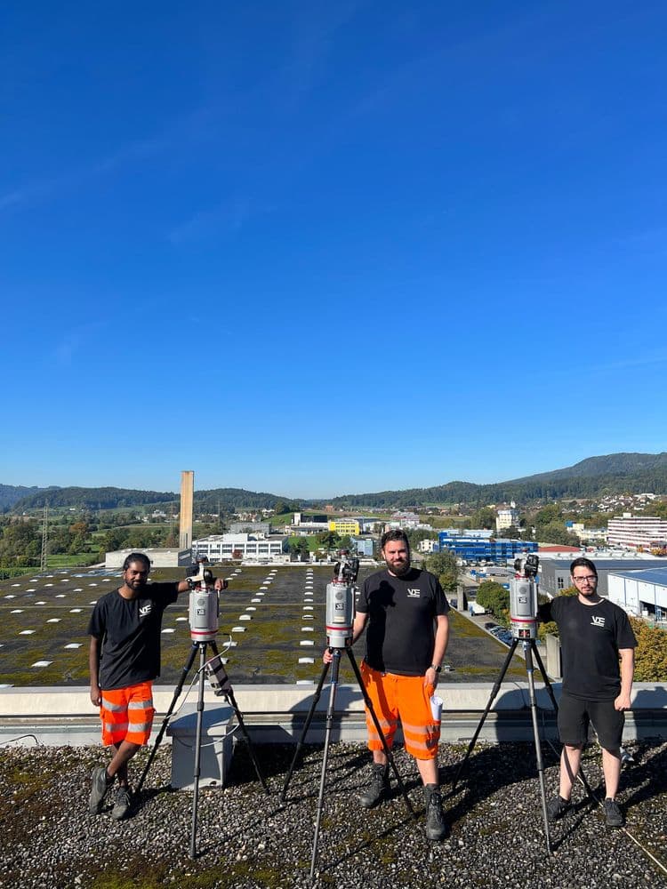 Drei Vermessungstechniker mit Totalstationen auf einem Flachdach, Baustellenkleidung in Orange, Industrie- und Stadtblick mit Hügeln im Hintergrund unter klarem blauem Himmel.