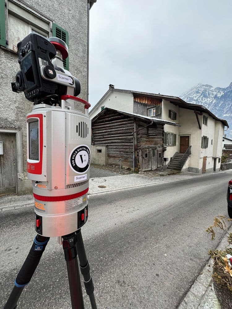 Straßenszene mit einem 3D-Laser-Scanner auf einem Stativ vor einem traditionellen Haus in den Bergen.