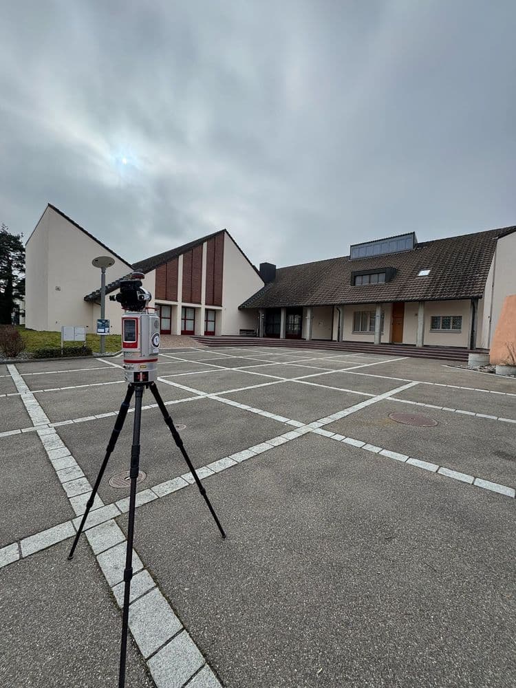 Gebäude mit Parkplatz, Himmelsbewölkung, und im Vordergrund ein Vermessungsgerät auf einem Stativ.