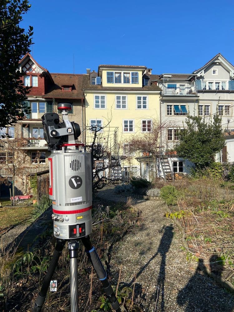 Ein Laserscanner steht im Vordergrund eines Gartens vor mehrstöckigen Wohnhäusern unter klarem Himmel.