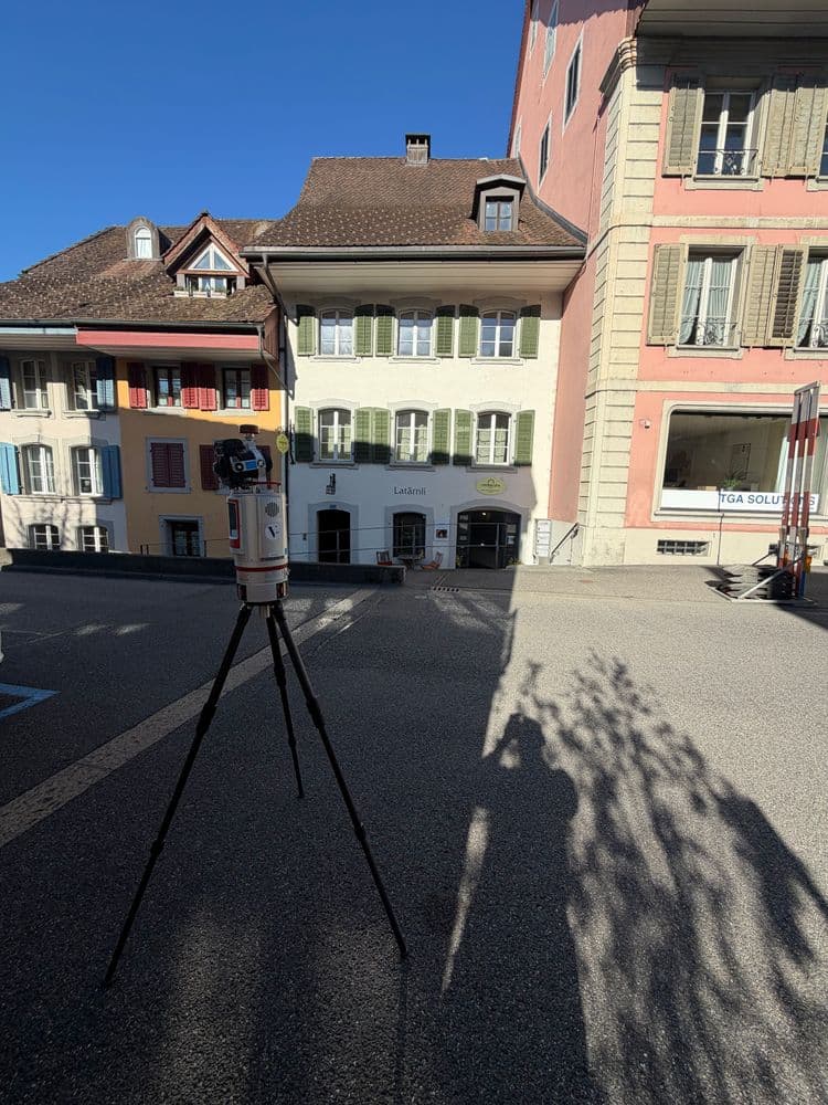 Stativ mit Vermessungsgerät auf einem Platz vor historischen Wohnhäusern mit Fensterläden, sonniger Himmel, lange Schatten und leere Straße in einer europäischen Altstadt.