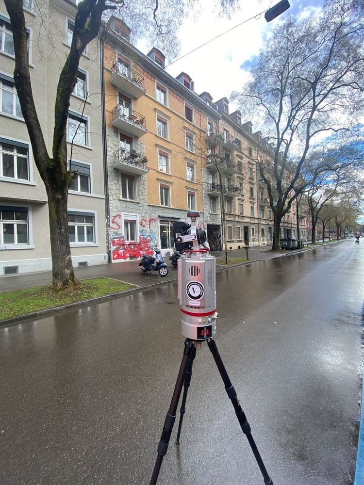 Vermessungsgerät auf Stativ in einer städtischen Straße mit mehrstöckigen Wohnhäusern im Hintergrund.