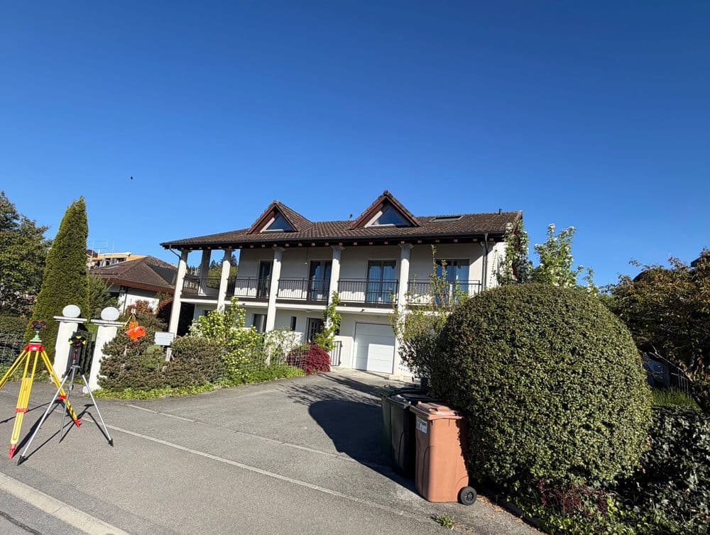 Zweistöckiges Wohnhaus mit Balkon und Satteldach, Einfahrt und Garage; vorne Vermessungsgeräte auf Stativ, Hecken und Büsche bei klarem blauem Himmel.