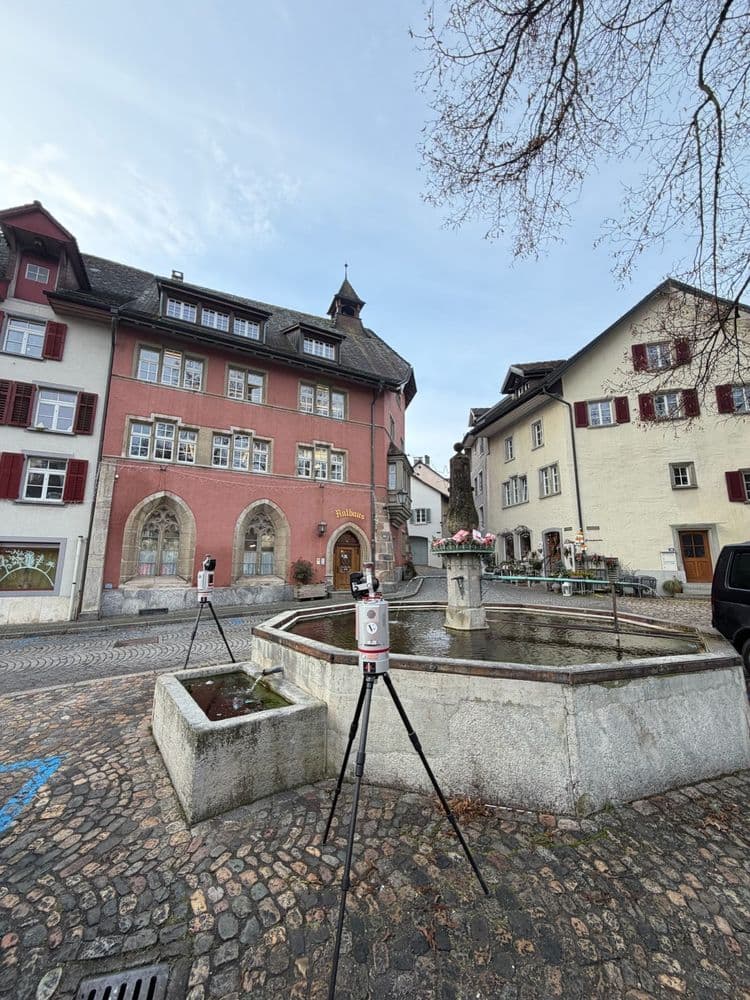 Historischer Marktplatz mit steinernem Brunnen und Stativ im Vordergrund, umgeben von alten Wohn- und Rathausgebäuden mit roten Fensterläden, Kopfsteinpflaster und blauem Himmel.
