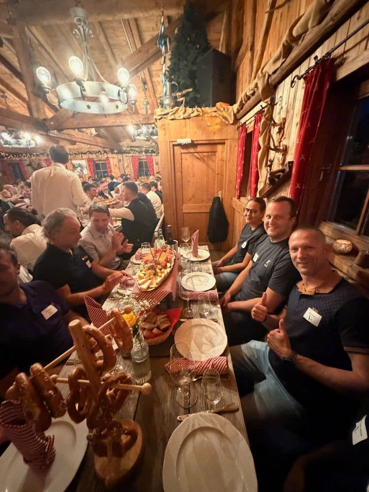 Gesellige Runde in rustikaler Berghütte: Männer sitzen an langer Tafel mit Brezeln, Bier und Speisen; Holzwände, rote Vorhänge und Kronleuchter sorgen für urige Stimmung.