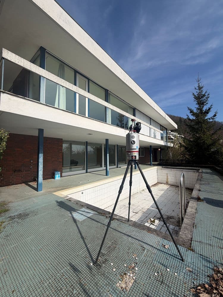 3D-Laserscanner auf Stativ vor modernem Gebäude mit Balkon und großen Fenstern; Vermessung eines leeren, gefliesten Schwimmbeckens im Außenbereich bei blauem Himmel.