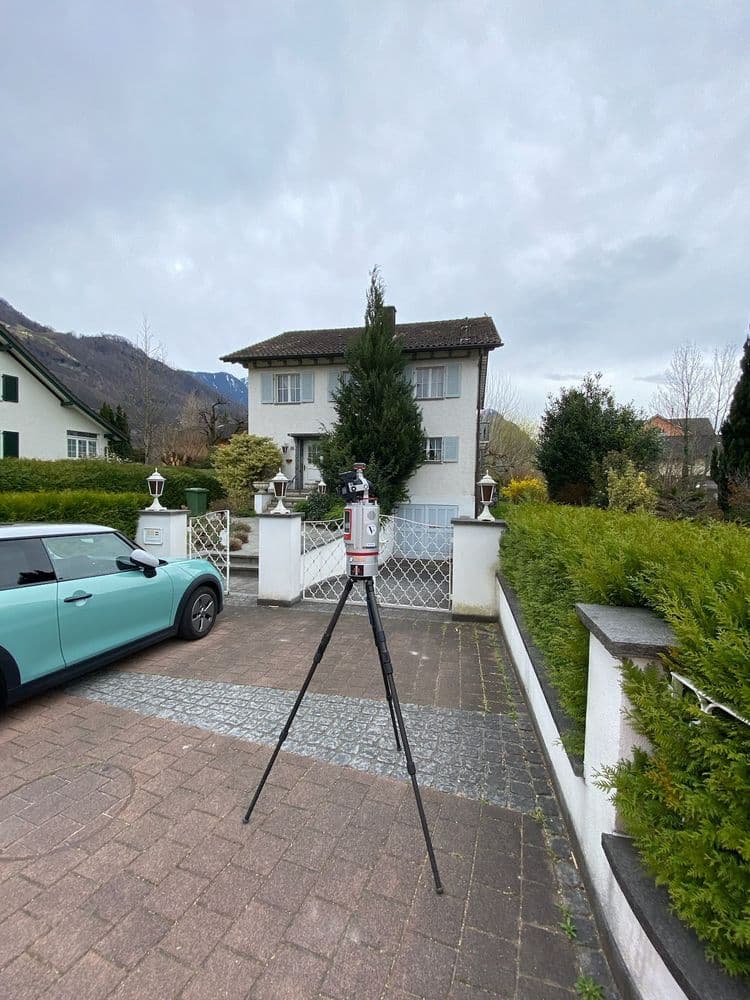 Ein weißes Haus mit einem dunklen Dach, umgeben von grüner Hecke und Bäumen. Im Vordergrund steht ein Stativ mit einem Vermessungsgerät auf einem gepflasterten Weg. Ein hellblaues Auto parkt links.