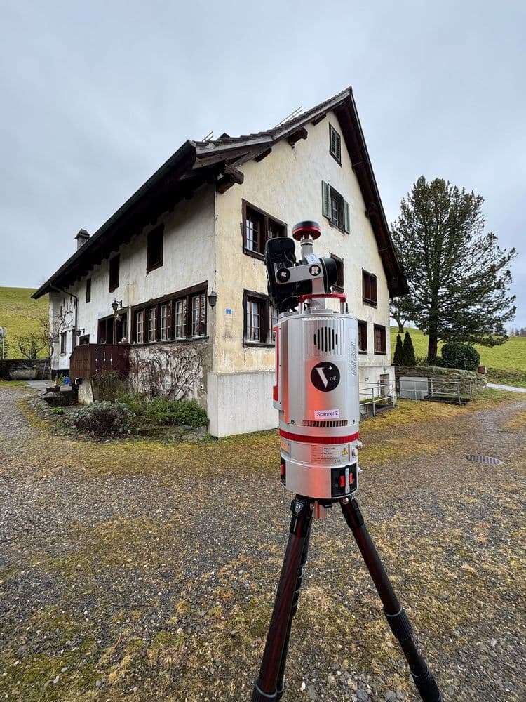 Eine Totalstation steht vor einem traditionellen, zweistöckigen Haus mit Schindeldach in ländlicher Umgebung.
