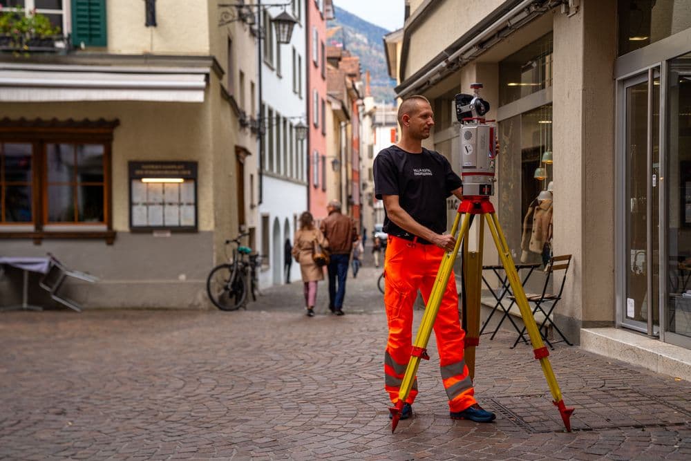 Vermessungsingenieur in Warnschutzkleidung arbeitet mit einem Stativ-Scanner in einer Kopfsteinpflaster-Gasse der Altstadt; Passanten und Fahrräder im Hintergrund, Berge in der Ferne.