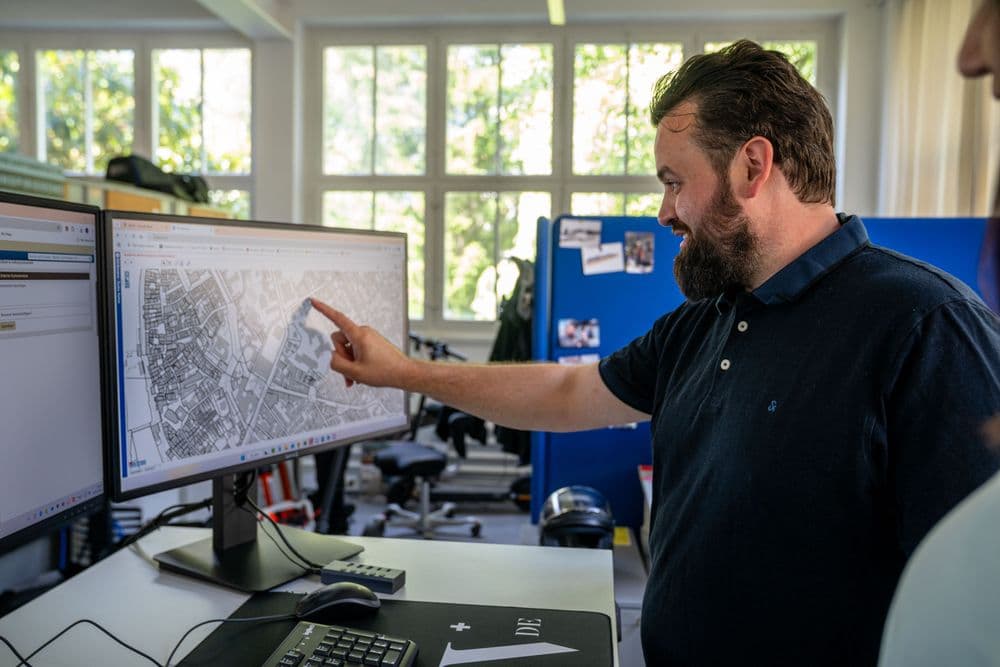 Mann im Büro zeigt auf Stadtplan auf großem Monitor am Schreibtisch, Computerarbeitsplatz mit Tastatur und Büroausstattung, Tageslicht durch große Fenster.