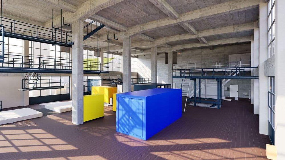 Helle Industriehalle mit Betonstützen, Galerien und Treppen; im Vordergrund große blaue und gelbe Container/Boxen auf Fliesenboden, Sonnenlicht fällt durch hohe Fenster.