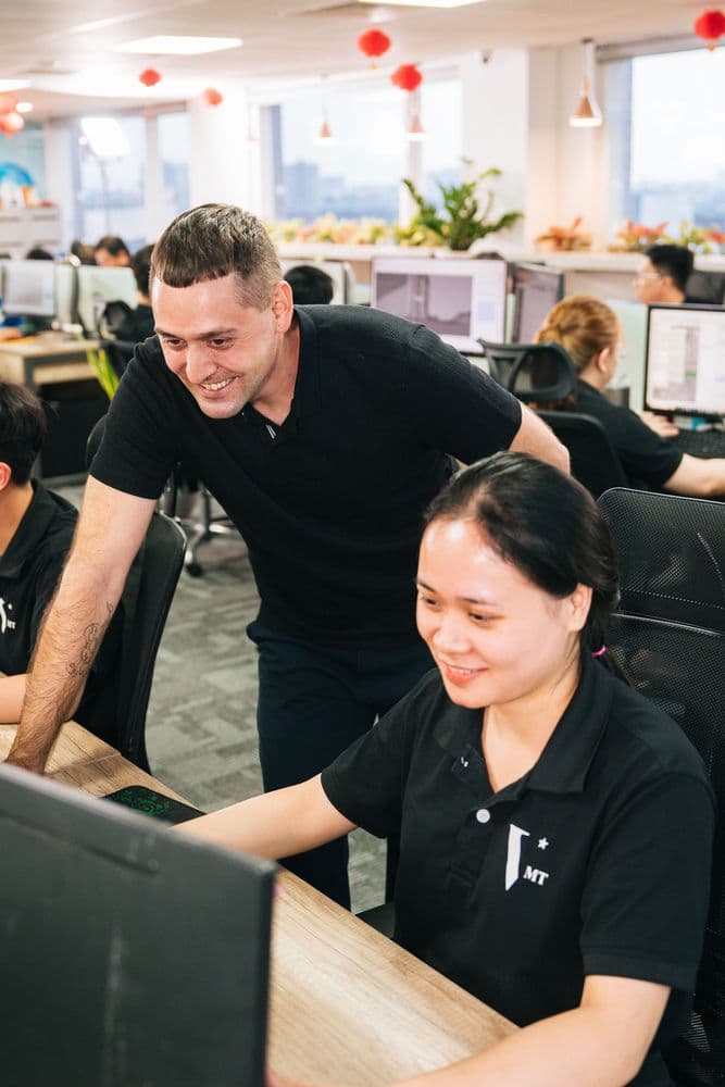 Mitarbeiter unterstützt Kollegin im modernen Großraumbüro am Computer, Teamarbeit im IT- oder Kundenservice-Umfeld mit mehreren Arbeitsplätzen und Monitoren im Hintergrund.