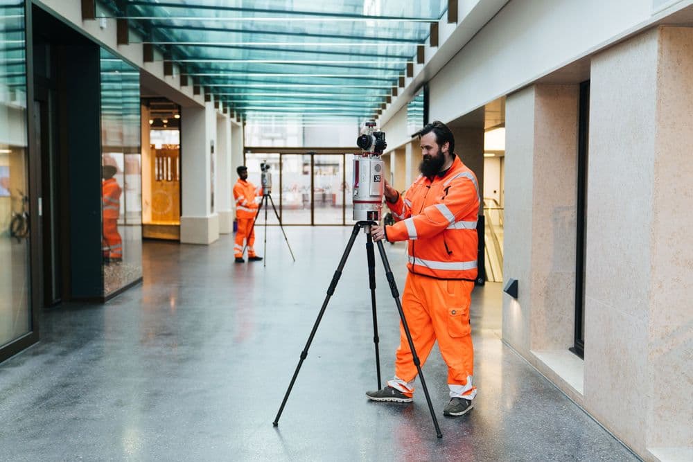 Vermessungstechniker mit orangefarbener Warnweste bedient einen Laserscanner auf Stativ in einer Altstadtstraße mit weißen Häuserfassaden und blauem Himmel.