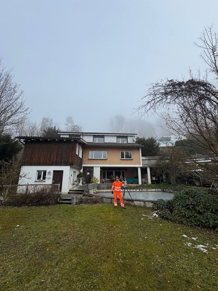 Mann in orangefarbener Arbeitskleidung steht im Garten vor einem zweistöckigen Haus mit Anbau, Rasen und kahlen Bäumen an einem nebligen Wintertag.