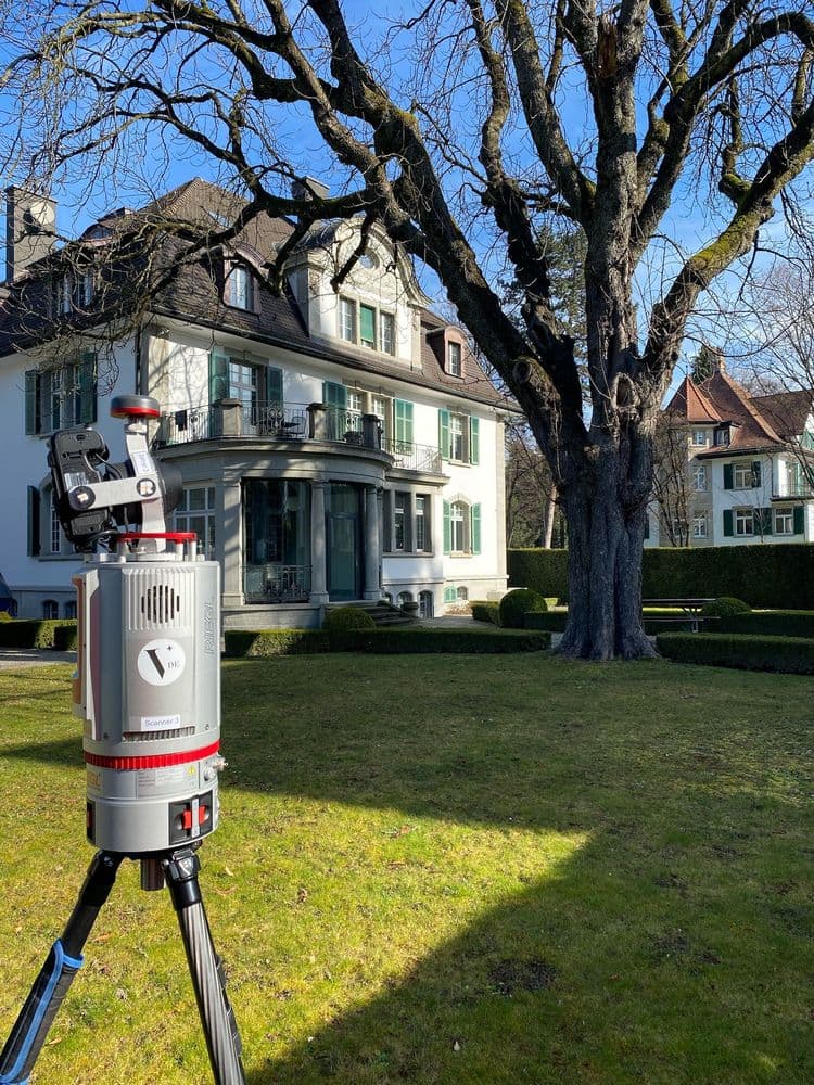 Eine historische Villa mit einem großen Baum im Vordergrund. Ein modernes Vermessungsgerät auf einem Stativ befindet sich auf dem Rasen vor dem Gebäude.