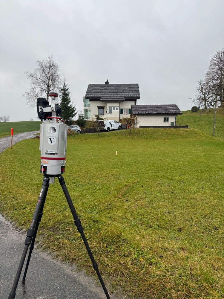 Ein Vermessungsgerät auf einem Stativ steht vor einem weißen Haus in ländlicher Umgebung.