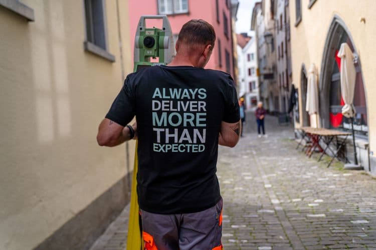 Ein Mann mit einem schwarzen T-Shirt, auf dem "Always deliver more than expected" steht, geht eine gepflasterte Gasse entlang.