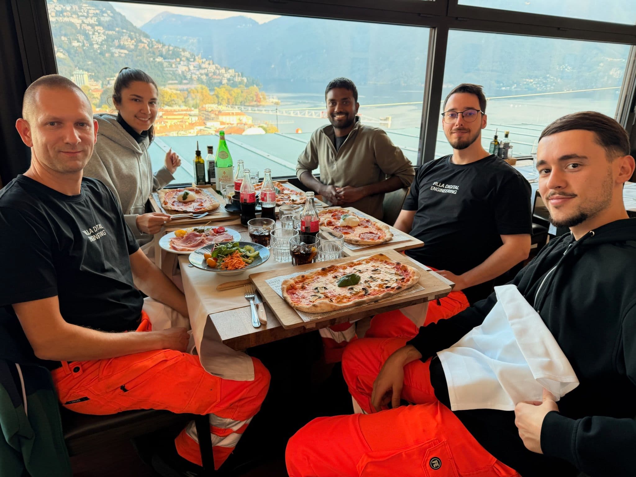 Gruppenfoto: Fünf Personen sitzen in einem Restaurant an einem Tisch mit Pizza, Salat und Getränken. Im Hintergrund ist eine malerische Aussicht auf Berge und Wasser zu sehen.