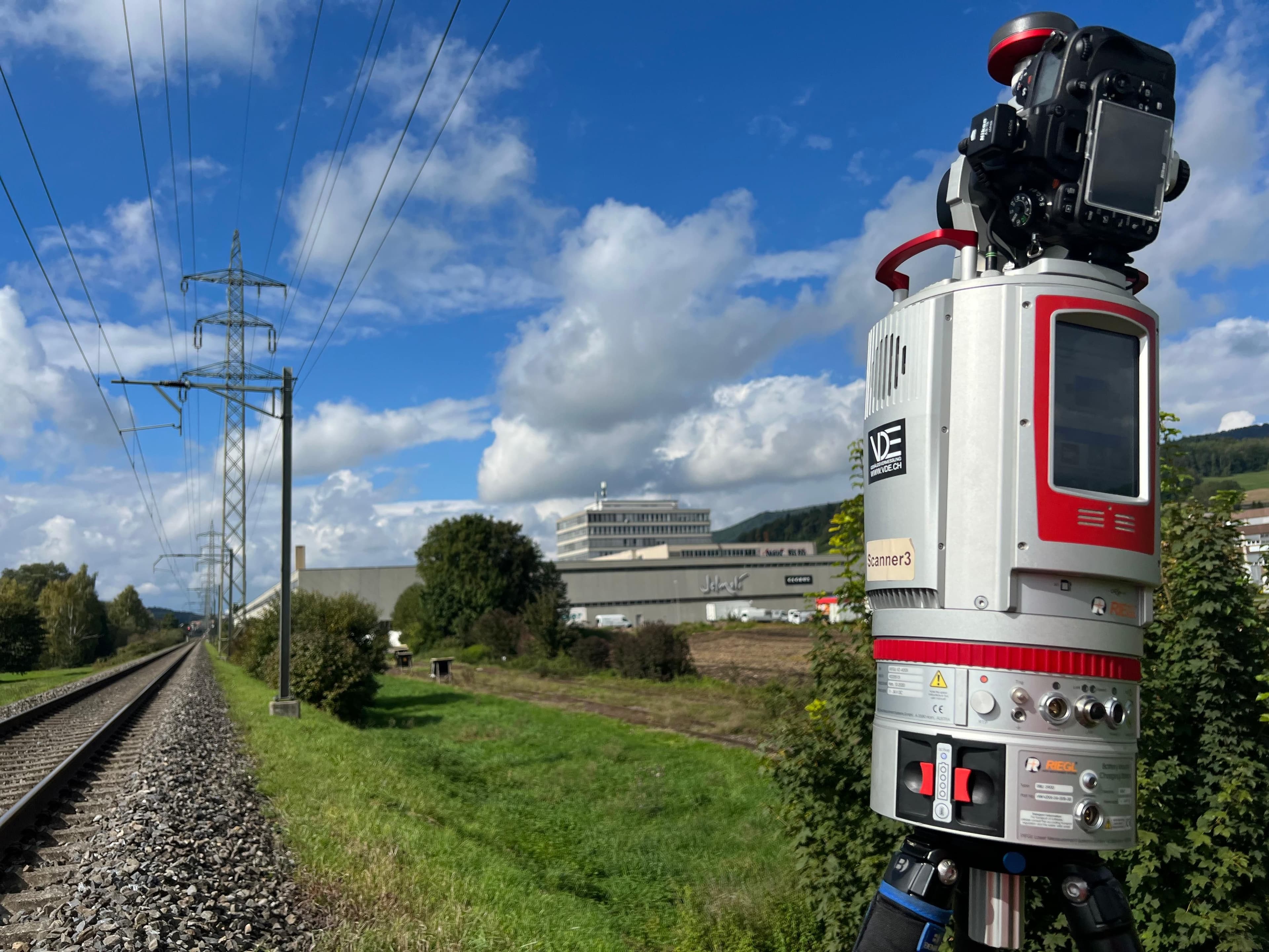 Industrievermessung: 3D-Laserscanner auf Stativ neben Bahnstrecke und Strommasten, mit Fabrikgebäude im Hintergrund unter blauem Himmel.