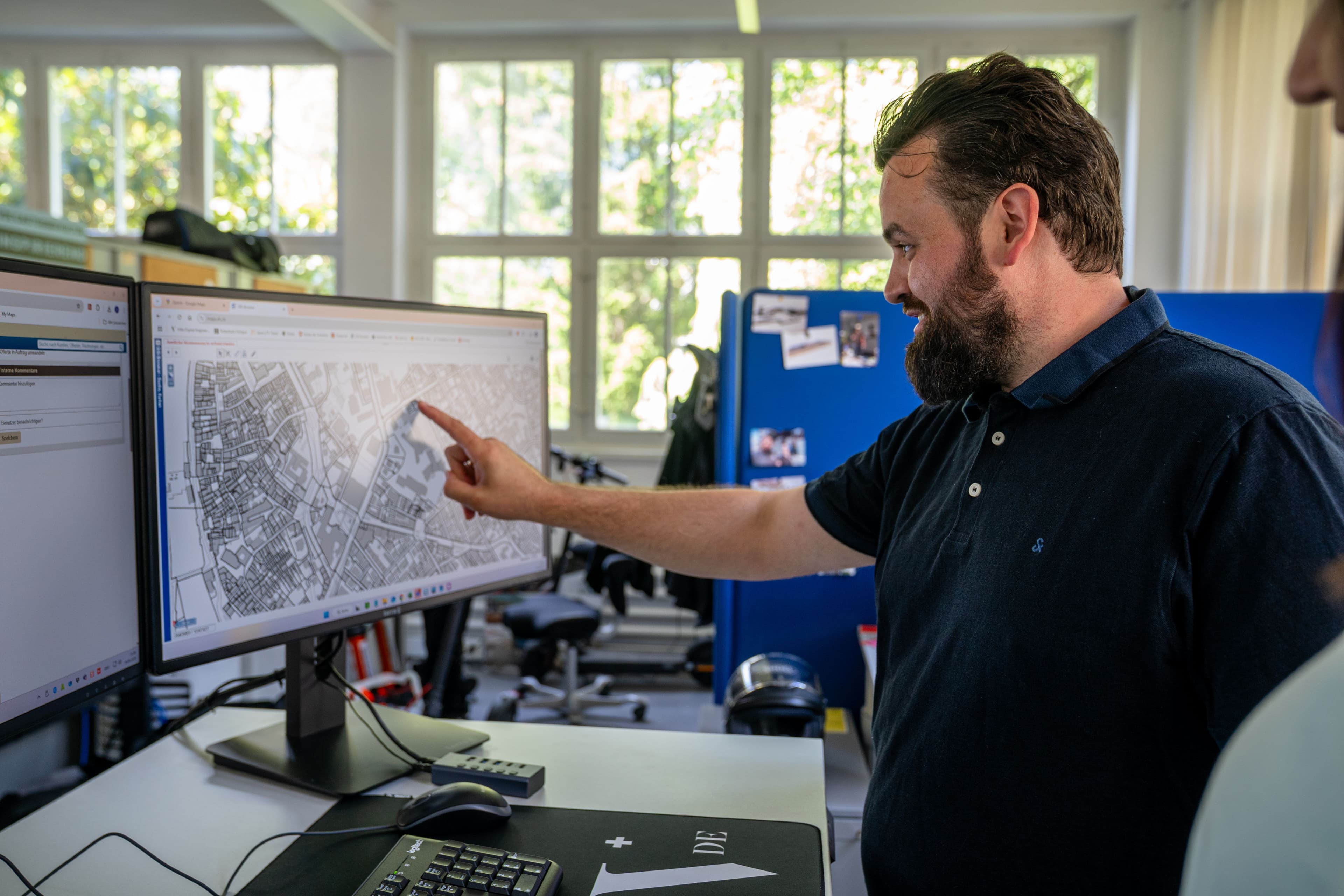 Mann im Büro zeigt auf Stadtplan auf großem Monitor am Schreibtisch, Computerarbeitsplatz mit Tastatur und Büroausstattung, Tageslicht durch große Fenster.