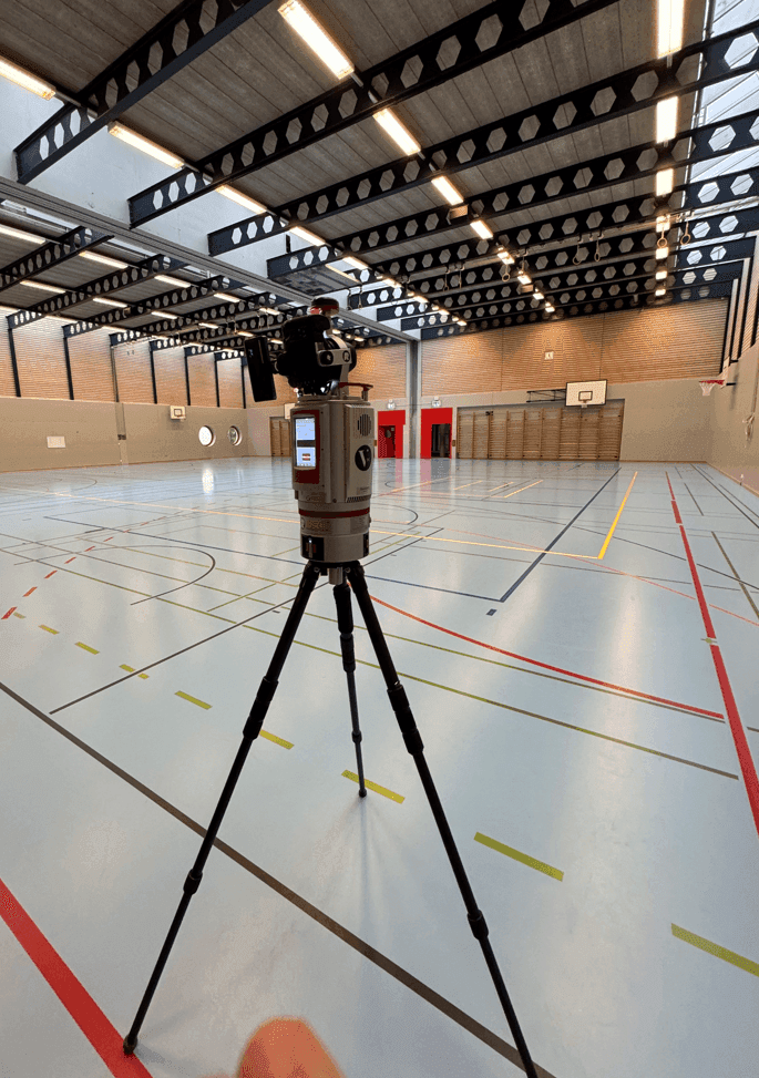 Eine geräumige Sporthalle mit Basketballkorb und einem aufgestellten Stativ mit Kamera im Vordergrund.